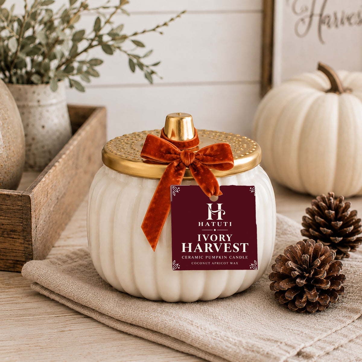 An ivory ceramic pumpkin candle with a ribbed white body, a gold lid, and a rust velvet bow, displayed on a linen cloth with pumpkins and pinecones in a styled setting. The front label reads 'HATUTI IVORY HARVEST'.