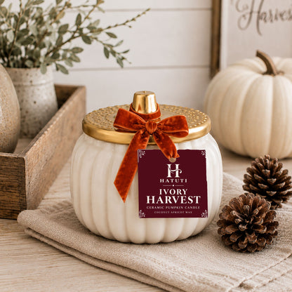 An ivory ceramic pumpkin candle with a ribbed white body, a gold lid, and a rust velvet bow, displayed on a linen cloth with pumpkins and pinecones in a styled setting. The front label reads 'HATUTI IVORY HARVEST'.